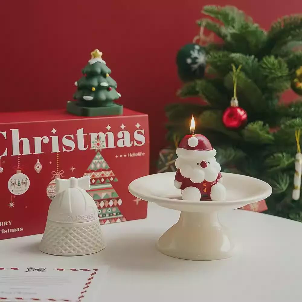 Decorative Christmas candle with Santa hat on a white stand, red 'Christmas' box, and Christmas tree in the background.