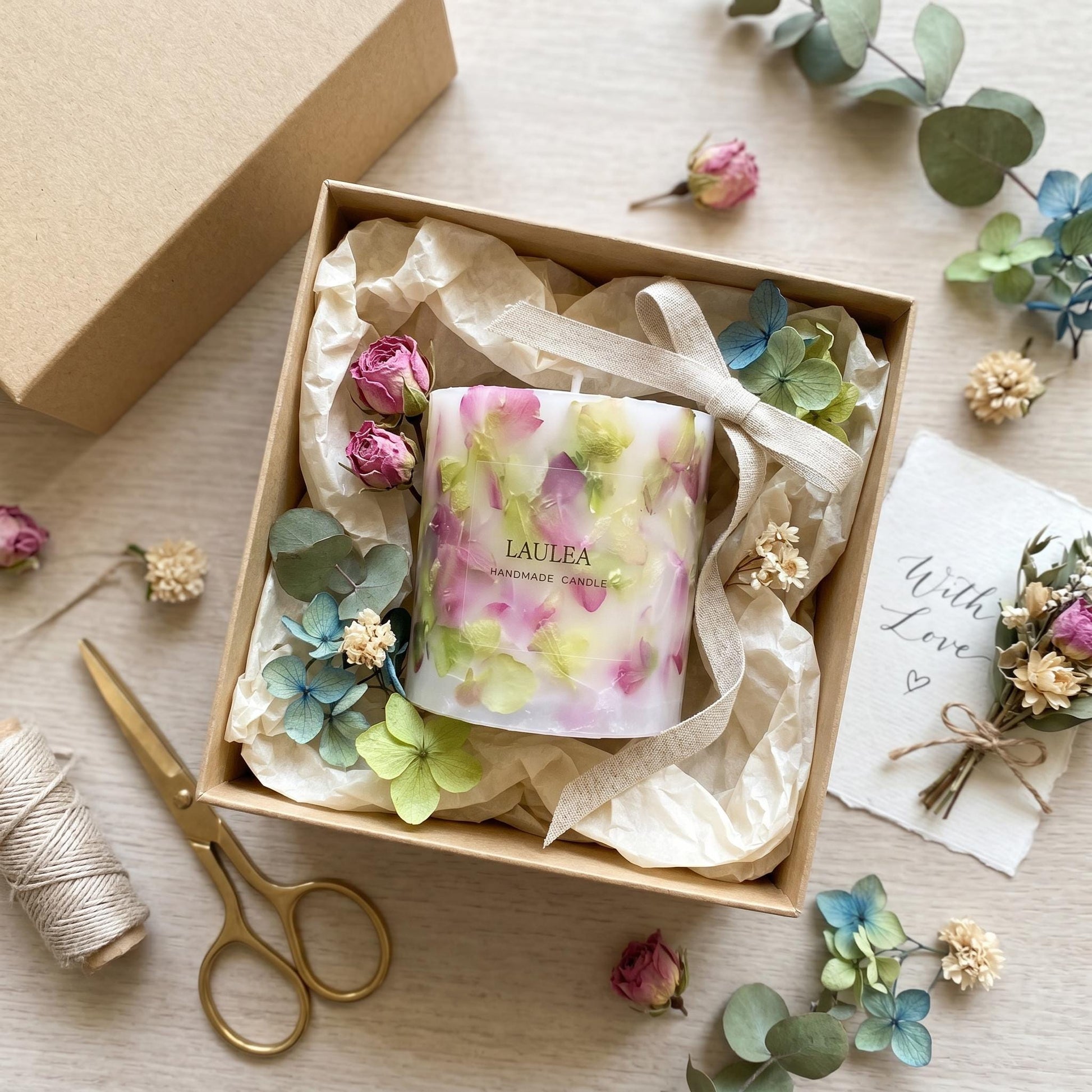 Decorative candle in a box with flowers and a card on a wooden surface