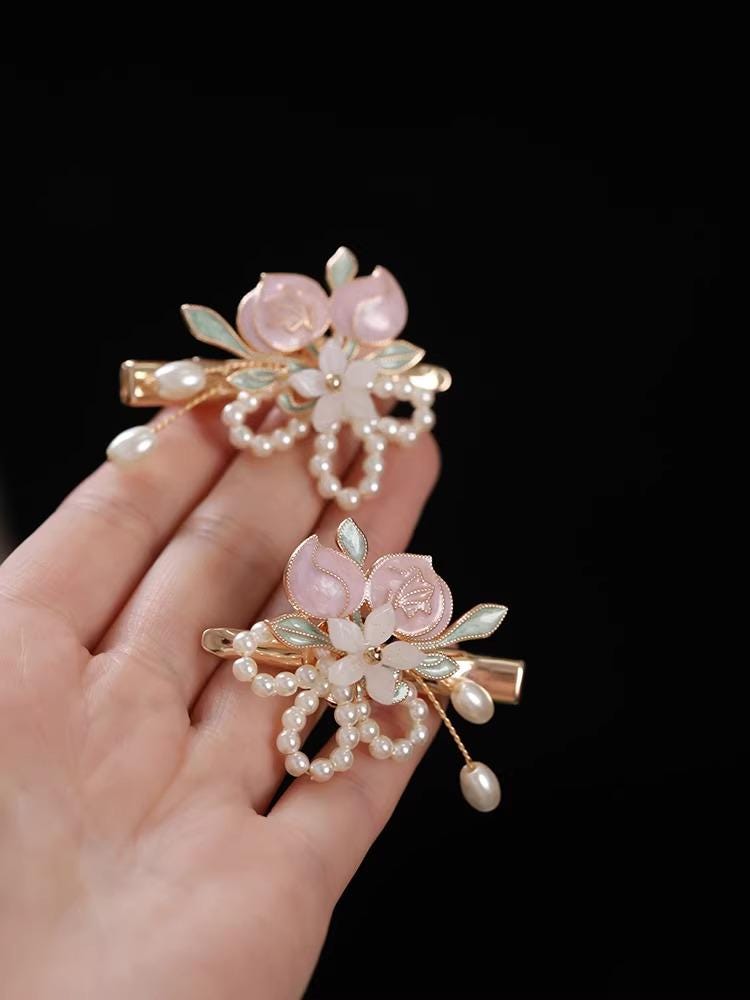 Decorative hair clips with floral and pearl details held by a hand against a black background