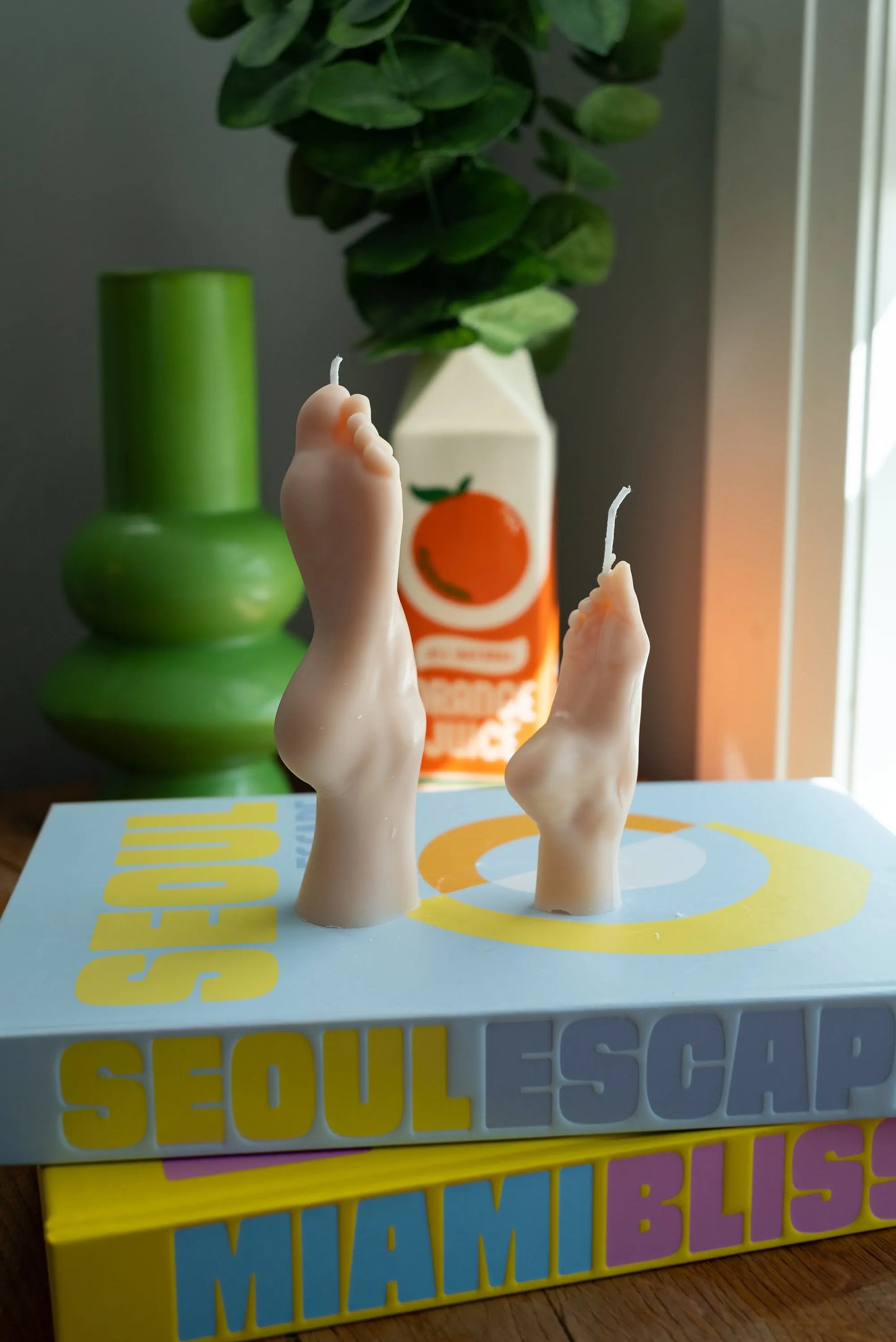 Candle with feet design on a book titled 'Seoul Escape Miami Bliss' with a green vase and orange juice box in the background.