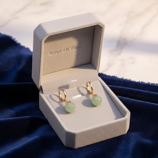 Gold earrings with jade stones in an open jewelry box labeled 'Scent Of Time' on a blue fabric background.