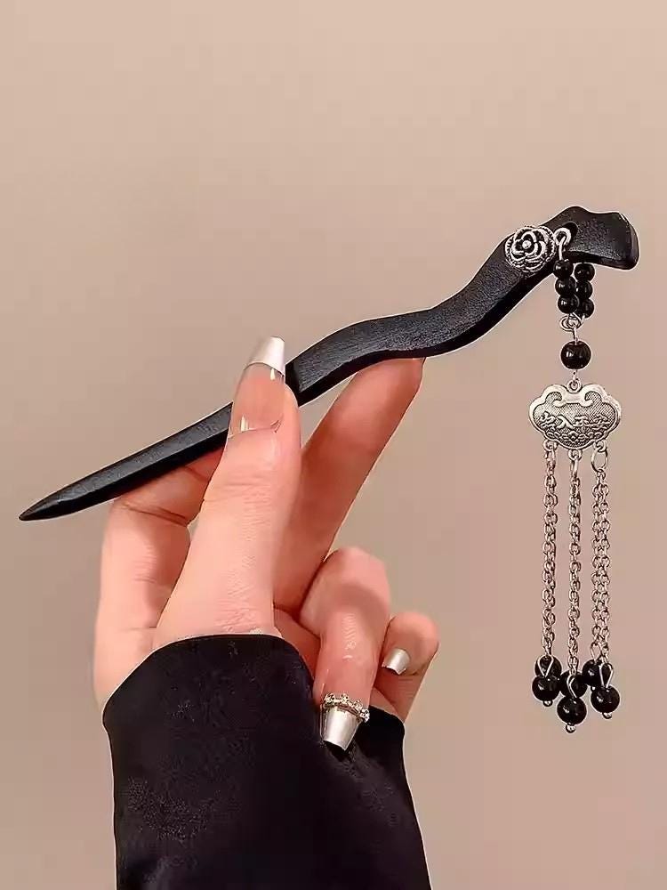 Hand holding a decorative hairpin with black hair clip and silver chain tassel against a beige background