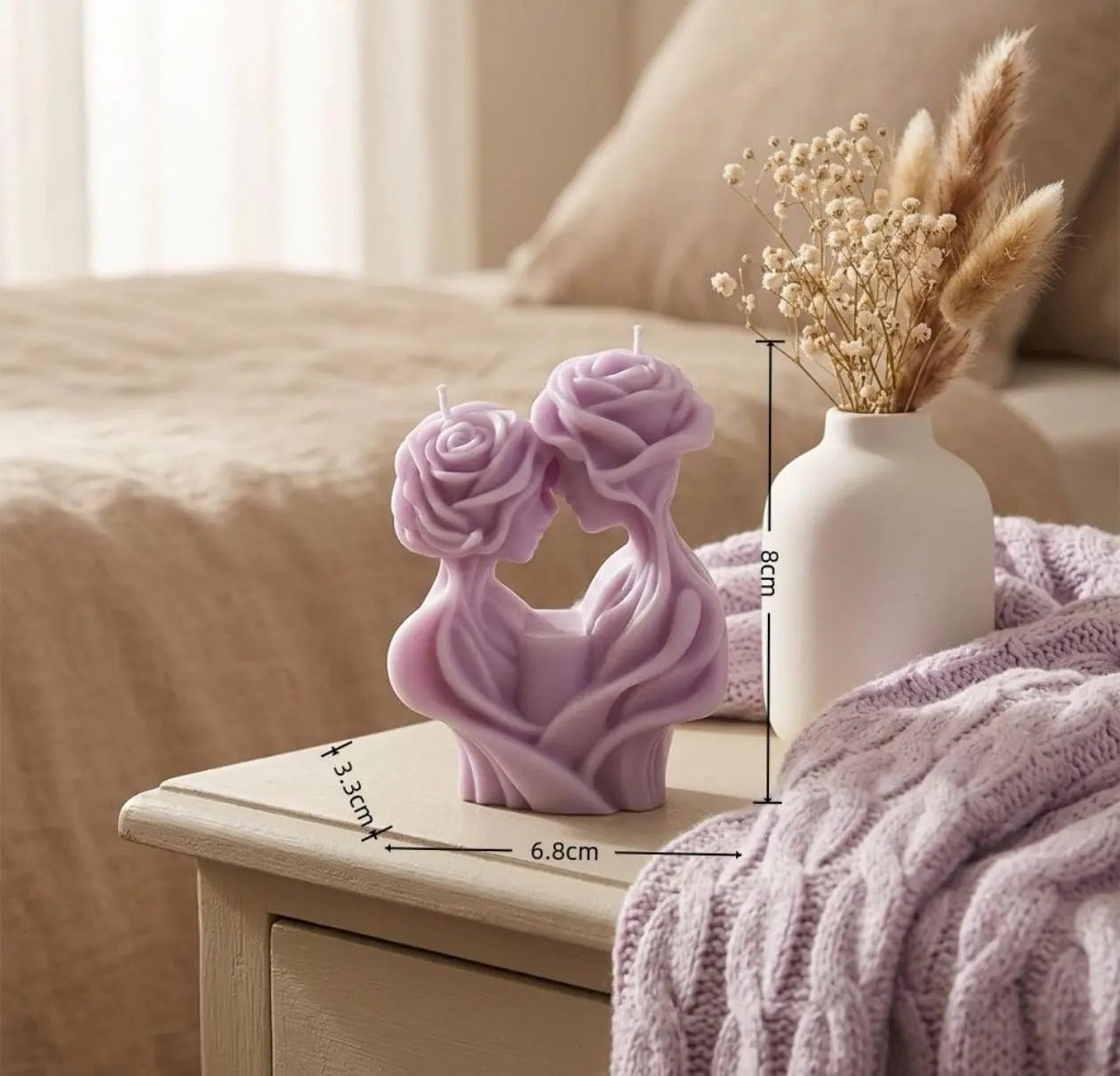 Decorative candles shaped like roses on a wooden surface with a vase of dried flowers in the background.
