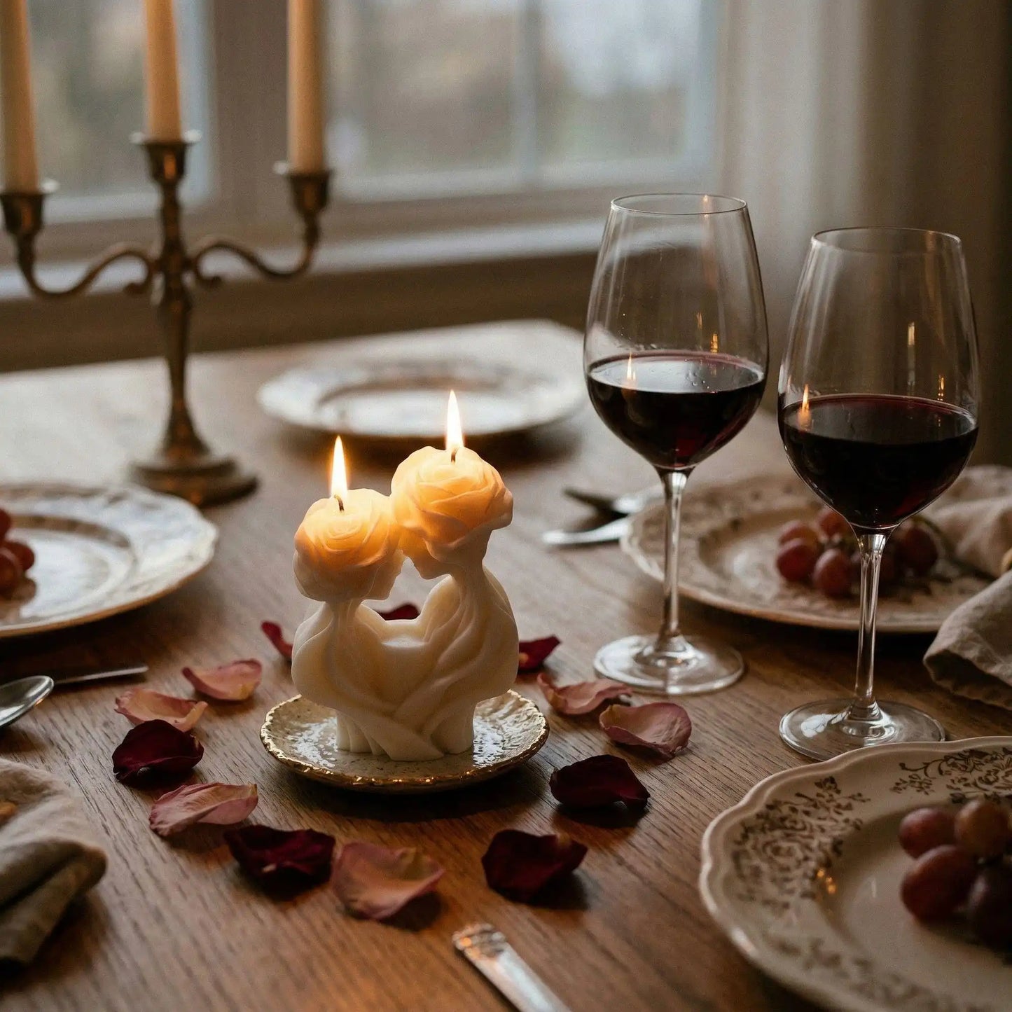 Decorative candles shaped like a couple on a table with wine glasses and rose petals.