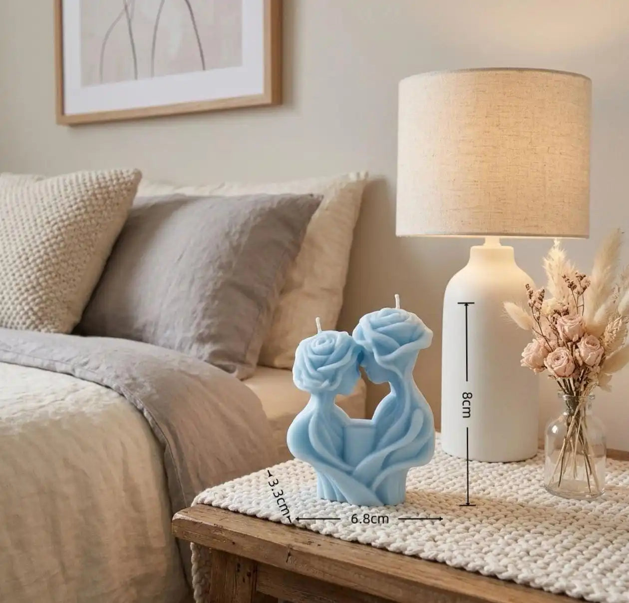 Decorative blue candle shaped like intertwined hearts on a bedside table with a lamp and vase in the background.