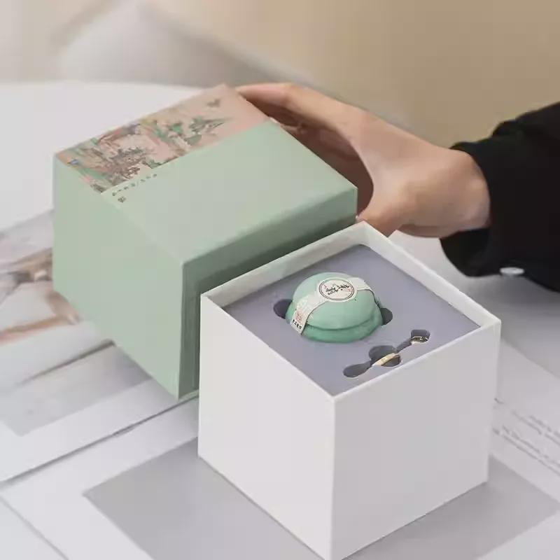 Solid Perfume in a white box with a hand reaching towards it, on a light surface.