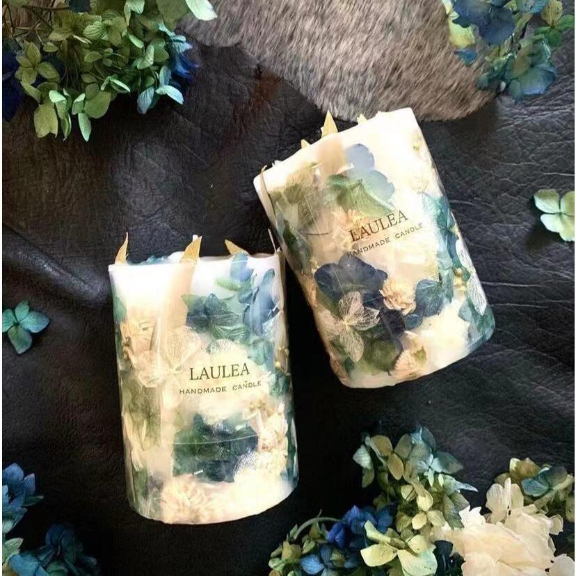 Two handmade candles with floral design packaging on a dark surface with greenery.