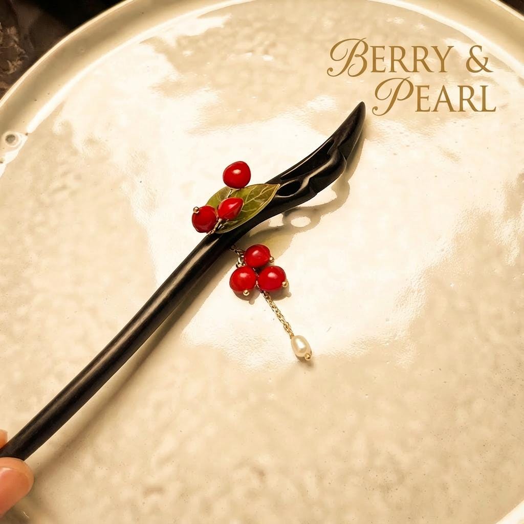 Black hairpin with red berries and a pearl on a textured beige surface, branded 'Berry & Pearl'.
