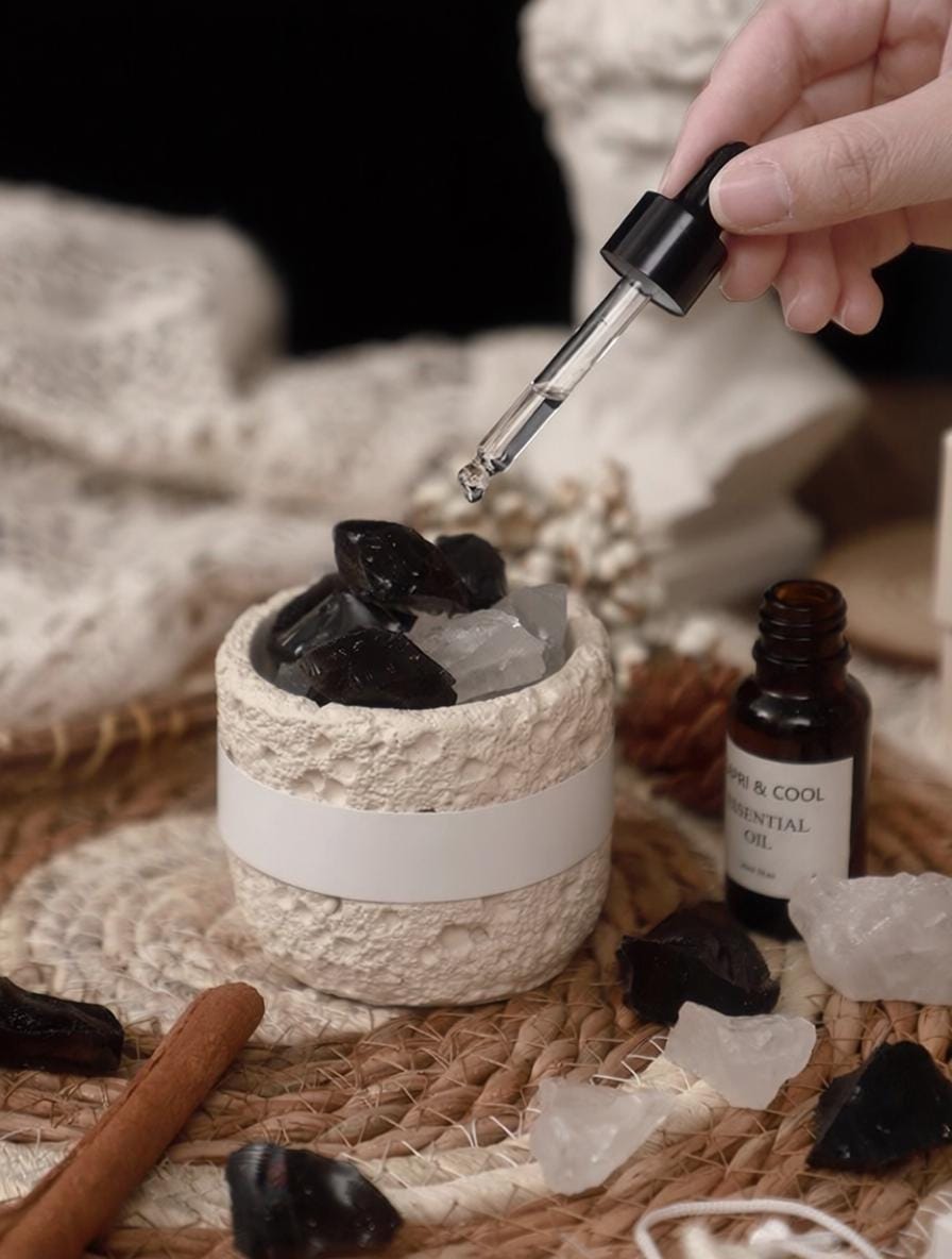 Hand dropping a liquid into a textured container with black stones, surrounded by essential oils and cinnamon sticks.