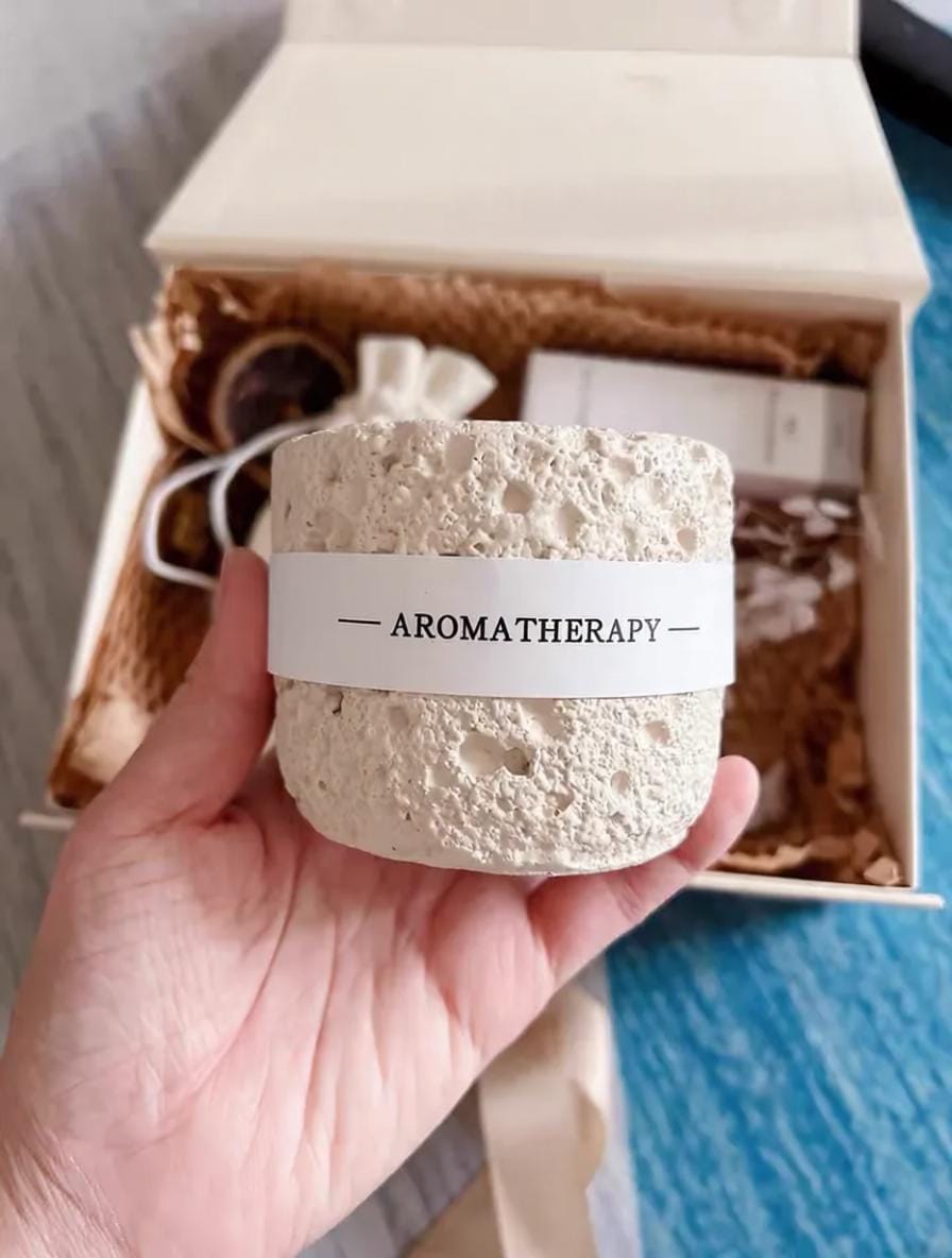 Hand holding a cream-colored container labeled 'Aromatherapy' in front of an open box.