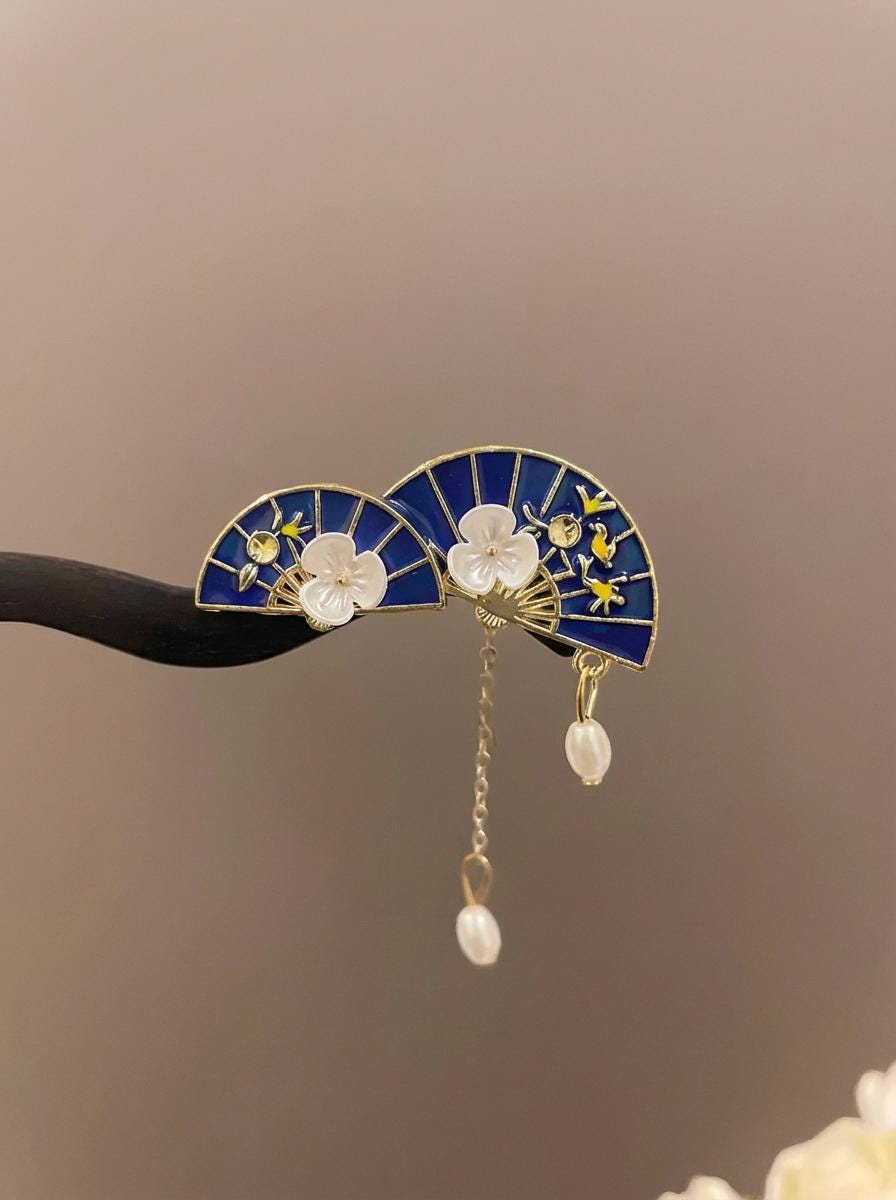 Decorative hair clip with blue fan design, white flowers, and gold accents on a blurred background.