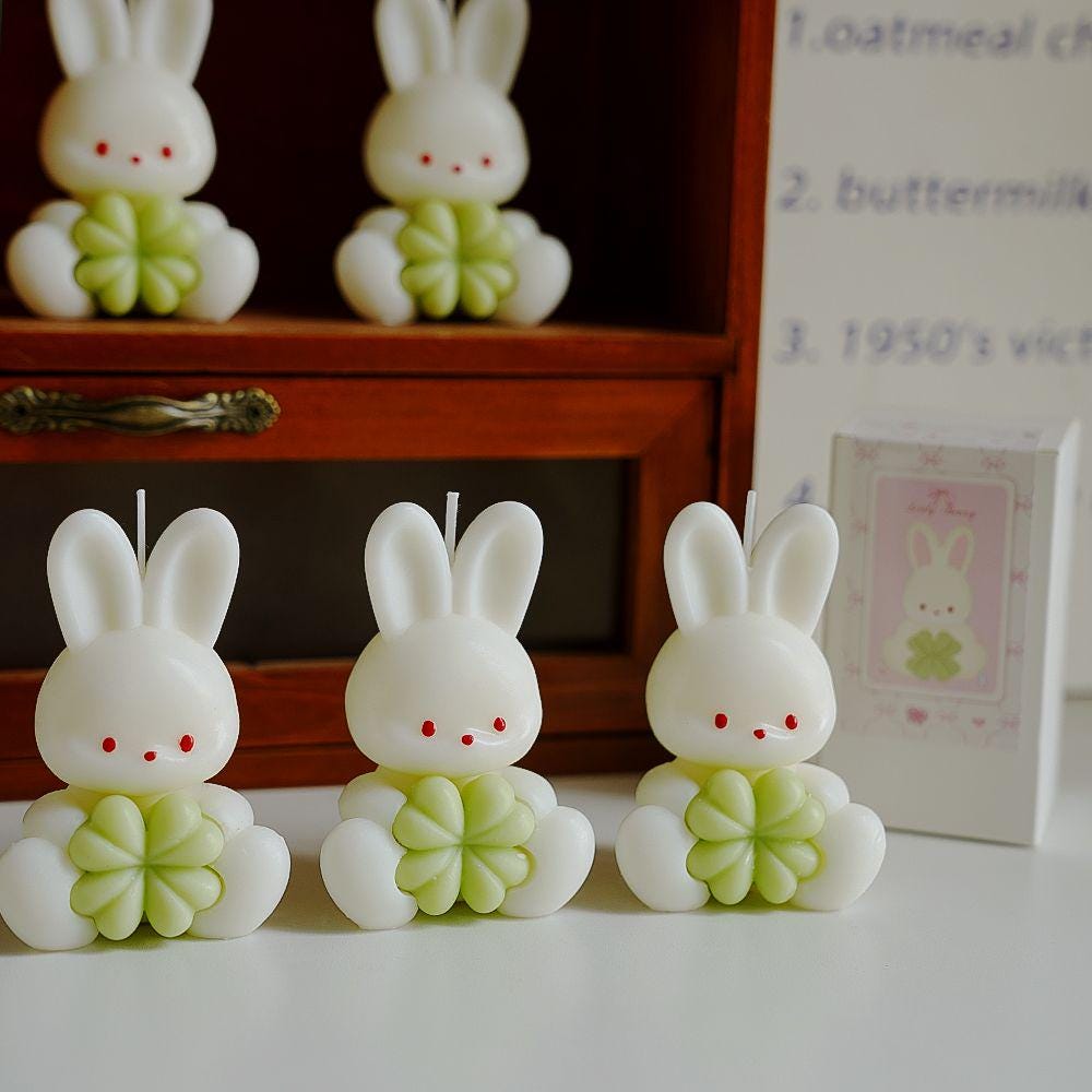 Three rabbit-shaped candles with green flowers on a white surface, with a wooden shelf and decorative items in the background.