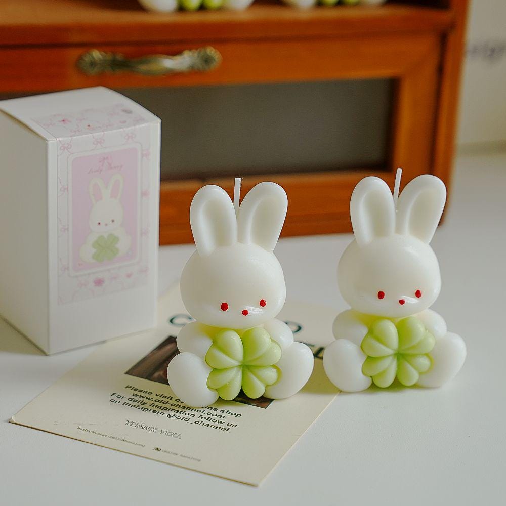 Two rabbit-shaped candles with green bodies on a table next to a box and paper.